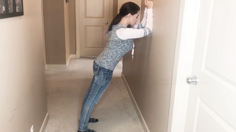 A woman performs a wall plank at home by leaning forward with her forearms against the wall and her body in a straight line