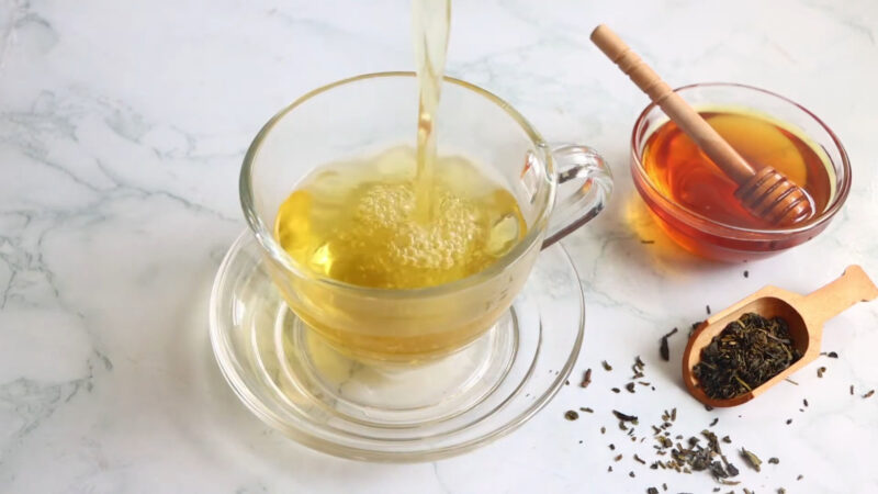 Glass cup filled with freshly brewed green tea, honey in a bowl with a wooden dipper, and a scoop of loose green tea leaves on a marble surface