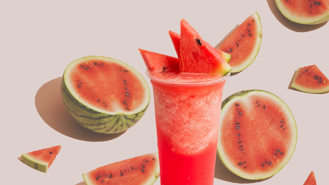A glass of watermelon juice garnished with watermelon slices, surrounded by halved and sliced watermelons
