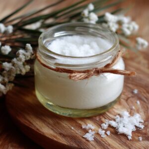 A small glass jar of homemade coconut oil deodorant tied with rustic twine, placed on a wooden board with coconut flakes scattered nearby