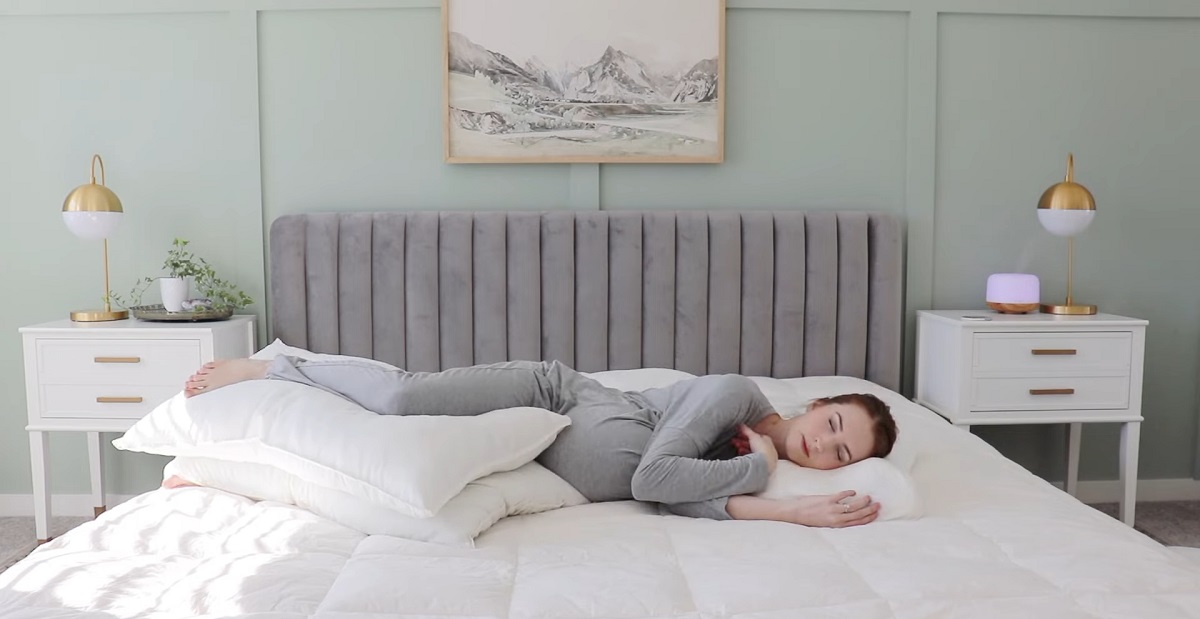 A woman in gray pajamas sleeps on her side in a cozy bedroom, using multiple pillows for support to maintain a comfortable resting position