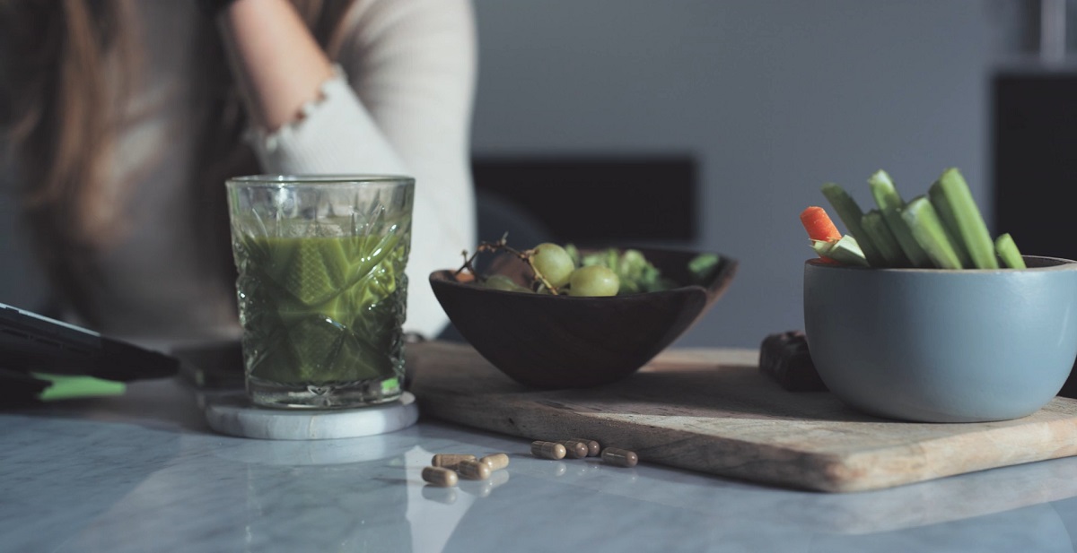 A glass of green juice alongside a bowl of grapes and a dish of celery and carrots on a wooden cutting board, with a person in the background