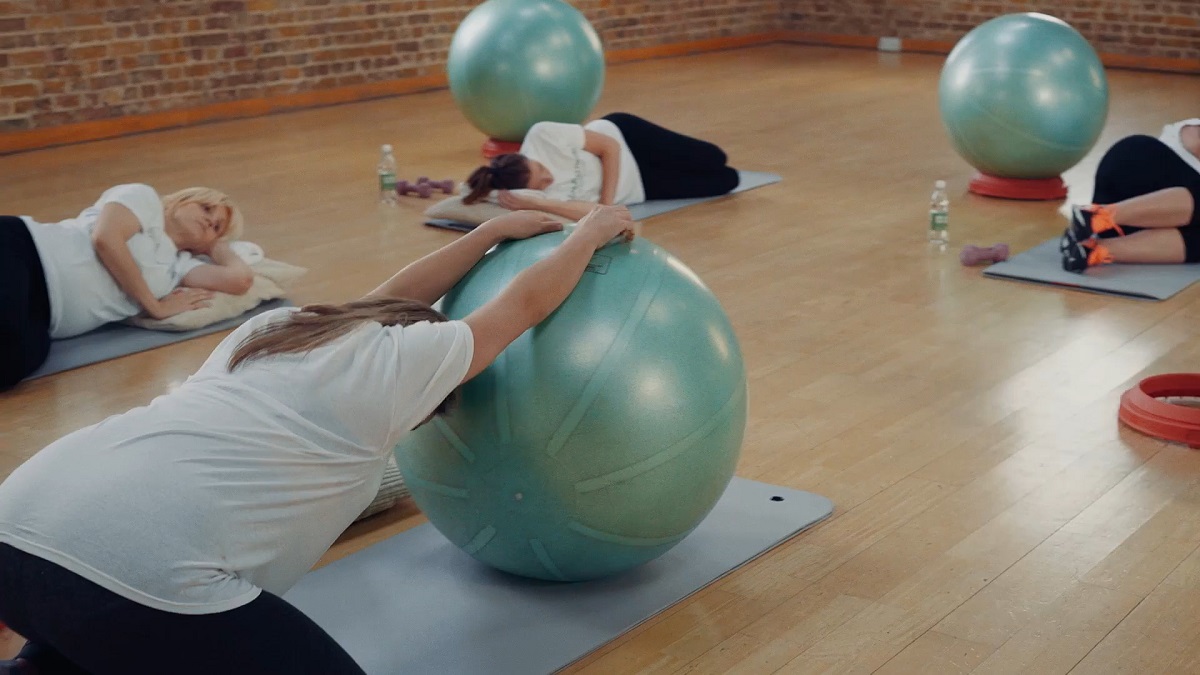 A group of pregnant women in a fitness studio perform prenatal exercises using large stability balls while lying on yoga mats