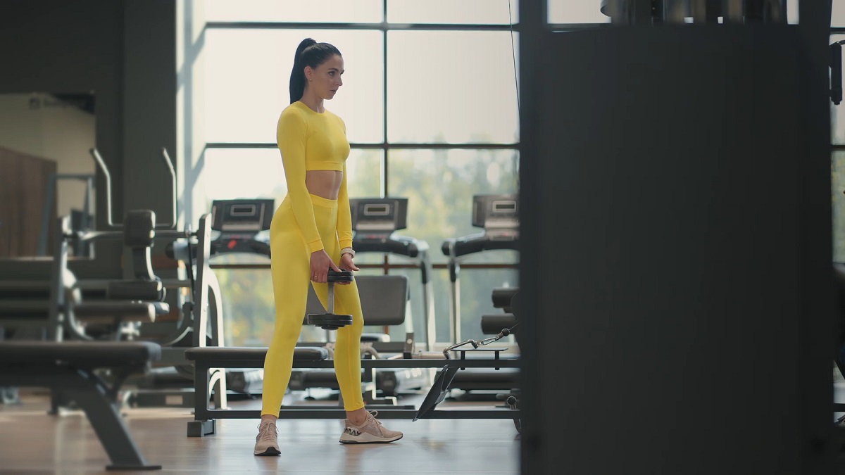 A woman in a bright yellow workout outfit lifting a dumbbell in a gym with treadmills in the background