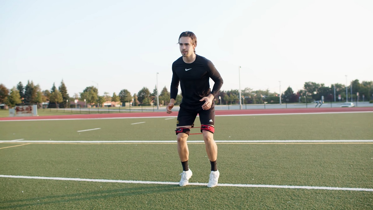 A male athlete in a black Nike outfit is performing agility drills on a green turf field with red resistance bands around his legs, working on speed and lateral movement