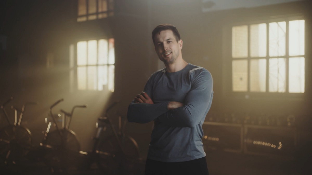 A confident and muscular man standing in a dimly lit fitness space with arms crossed, smiling at the camera