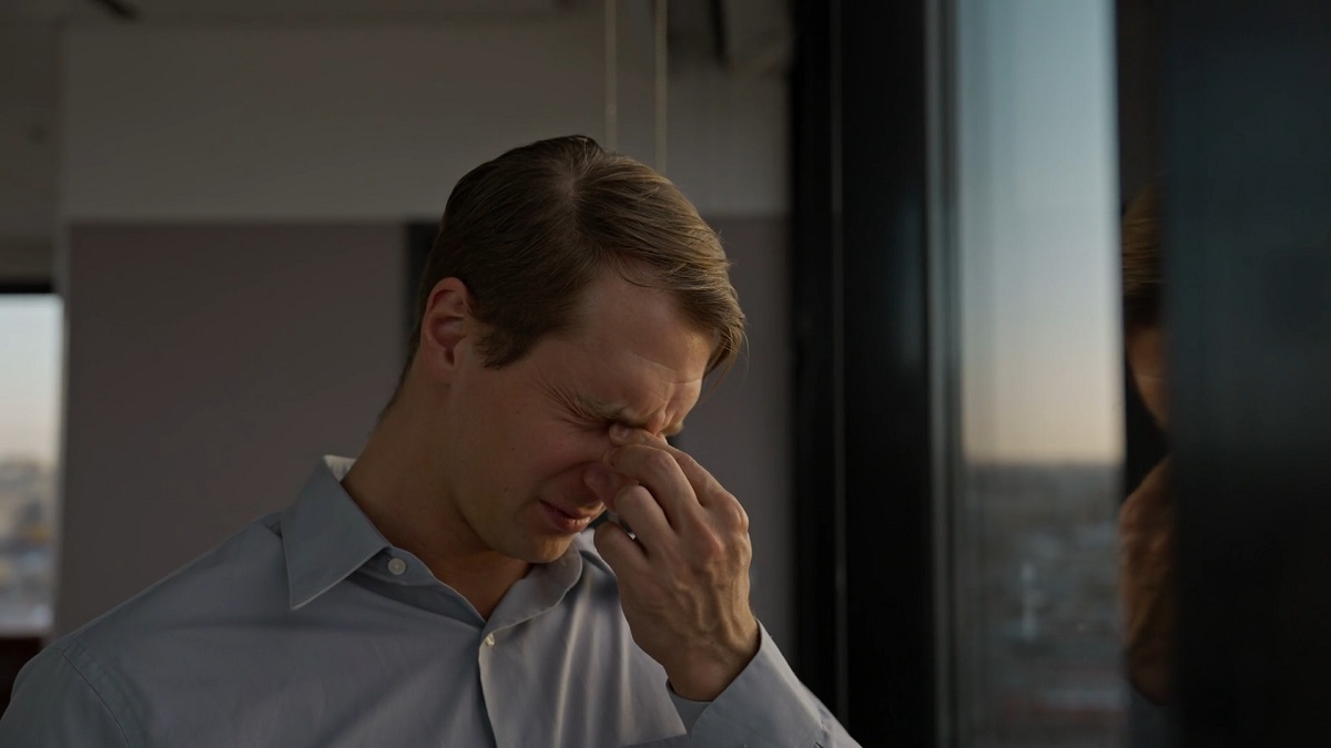 A man in a dress shirt stands by a window, rubbing his forehead with a pained expression, illustrating the cognitive symptoms of concussion