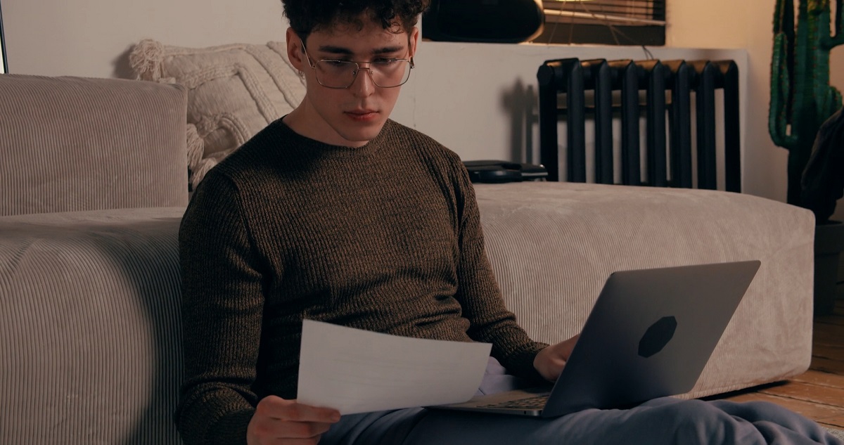 A young man with glasses sitting on the floor, reading a printed document while using a laptop