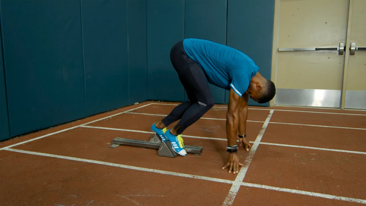 A male sprinter in a blue outfit and yellow track shoes is in the starting blocks on an indoor running track, preparing for an explosive sprint