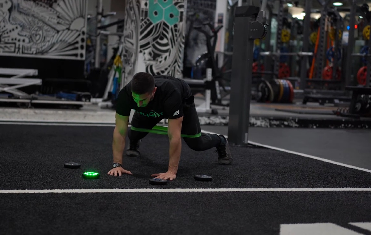 A man in a black athletic outfit is in a plank position, engaging with illuminated digital reaction pads on a black gym mat, training his reflexes and reaction time