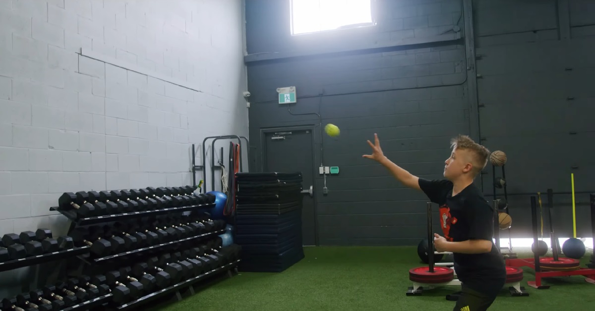 A young boy in a gym setting is seen tossing a tennis ball into the air, practicing hand-eye coordination