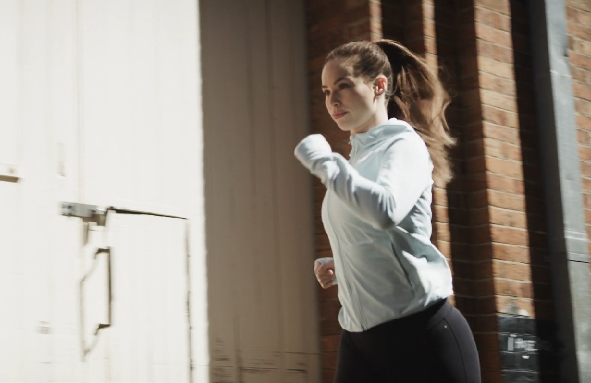 A woman jogging outdoors near a brick building, wearing a light blue workout hoodie, representing active lifestyle choices for fitness