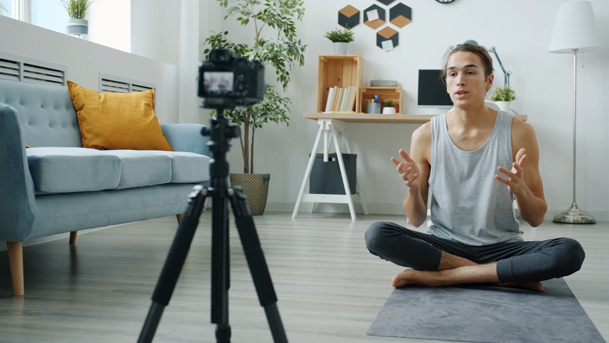 A young man in a gray tank top sits cross-legged on a yoga mat in a modern, well-lit living room, recording a video with a camera on a tripod