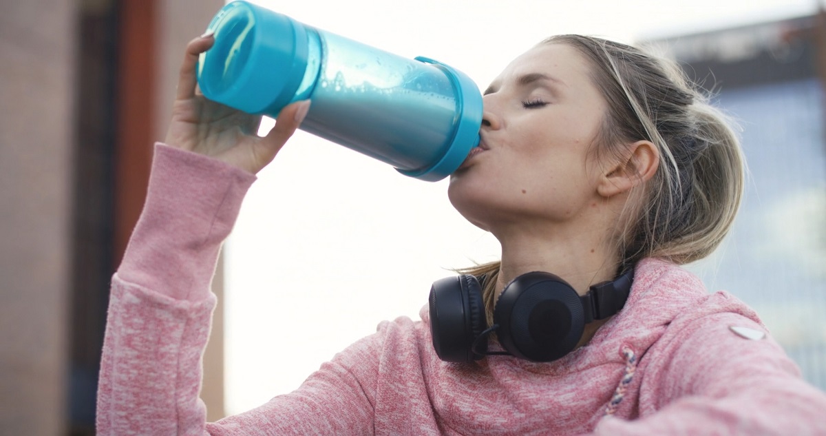 A woman in a pink hoodie drinking from a blue shaker bottle with black headphones around her neck