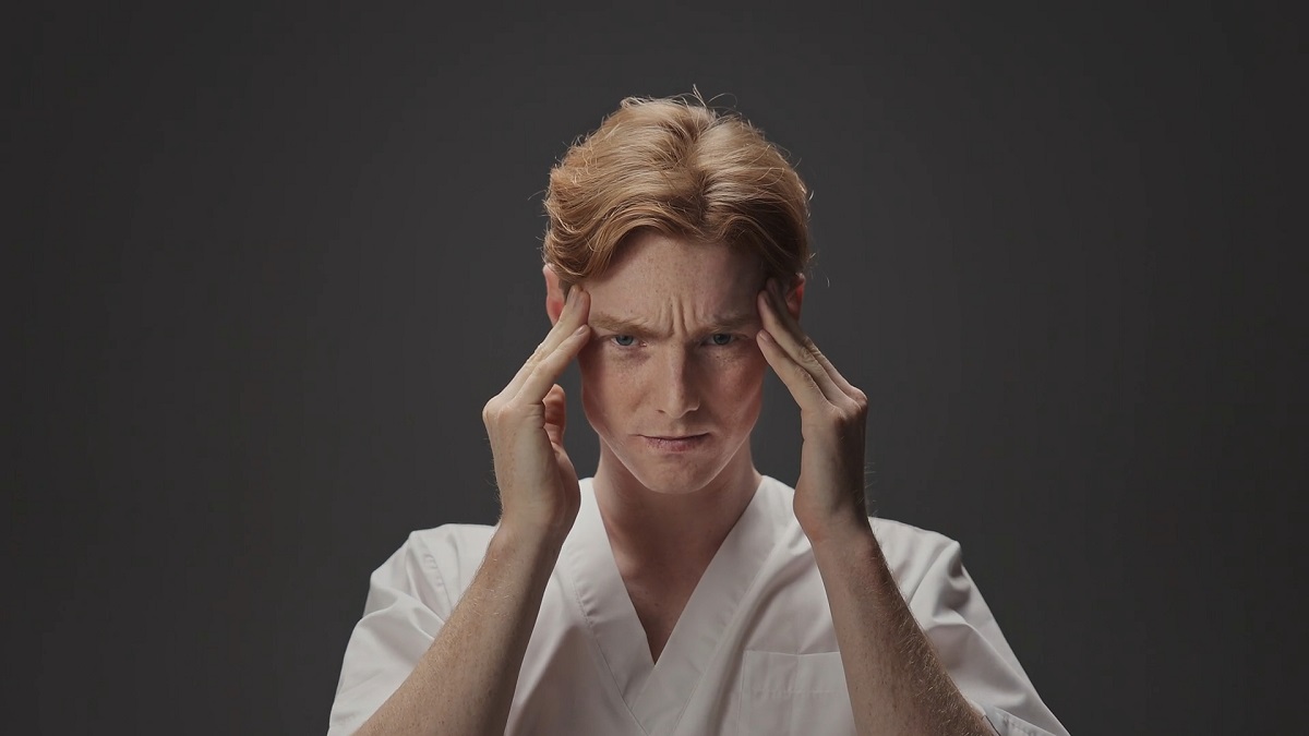 A concerned male doctor in a white coat presses his temples, indicating a headache or stress, a common symptom of concussion