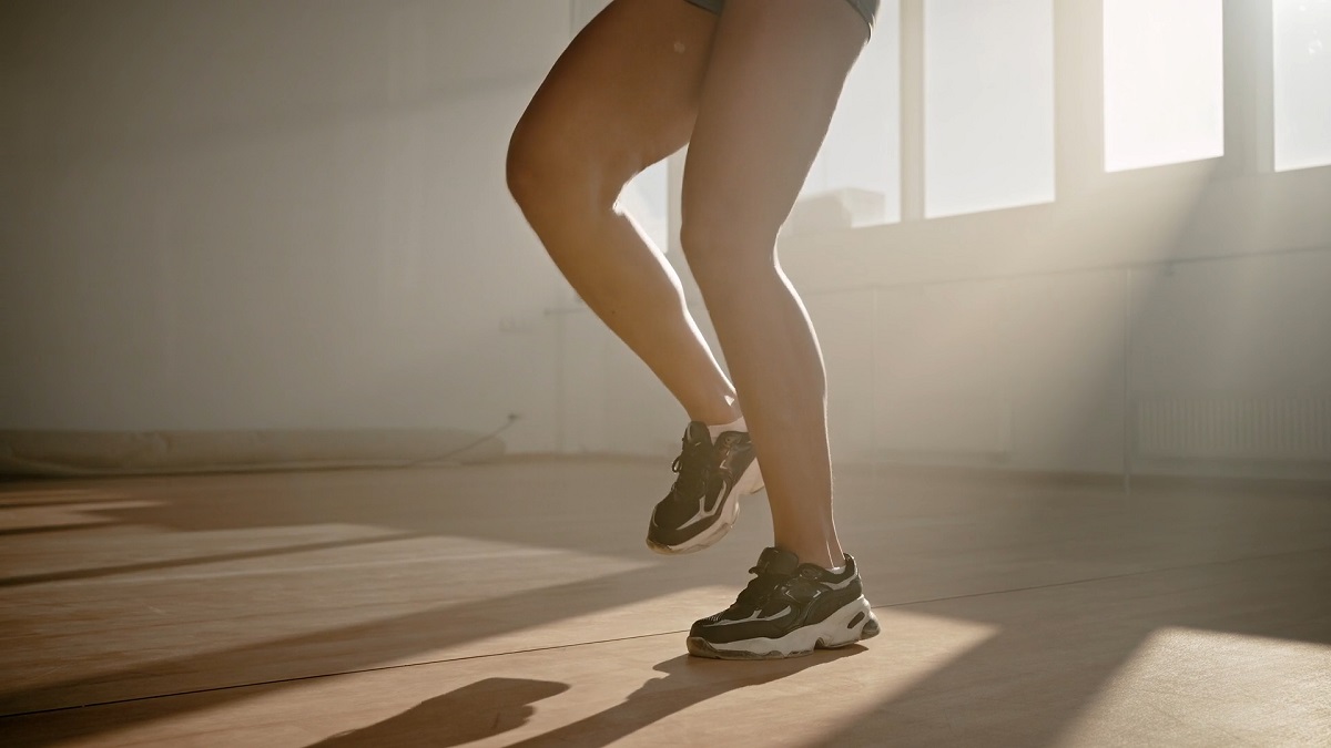 A close-up of a person's legs in black athletic shoes performing a marching or jogging-in-place movement in a well-lit gym