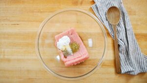 A glass bowl with raw ground turkey, yogurt, and spices on a wooden counter with a spoon and towel nearby