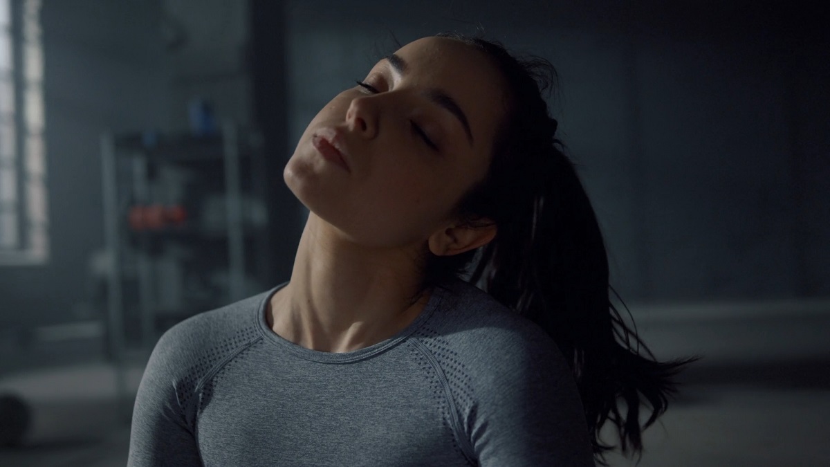 A woman in a gray long-sleeved athletic top tilts her head to the side in a dimly lit gym, stretching her neck muscles