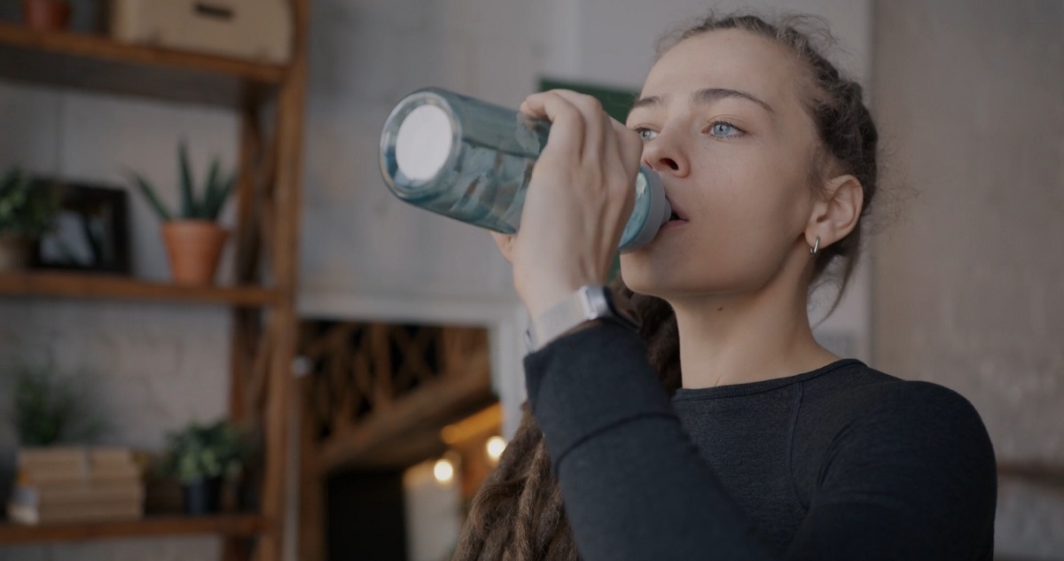 A young woman in athletic clothing drinks water from a bottle in a cozy indoor setting with plants