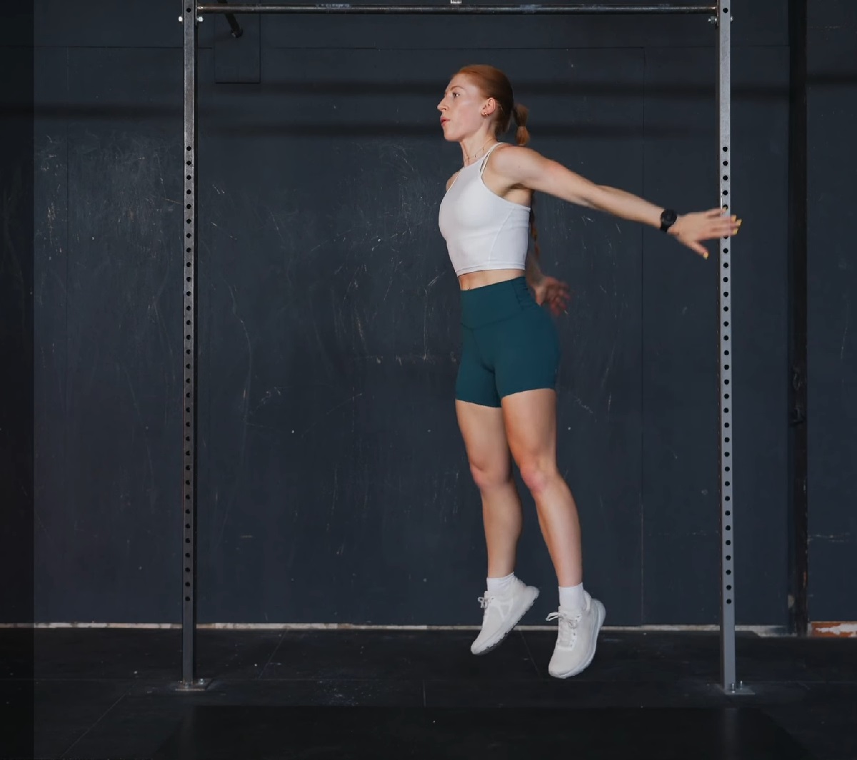 A fit woman in a white sports bra and teal shorts is mid-air, executing an explosive vertical jump against a dark background with a pull-up bar