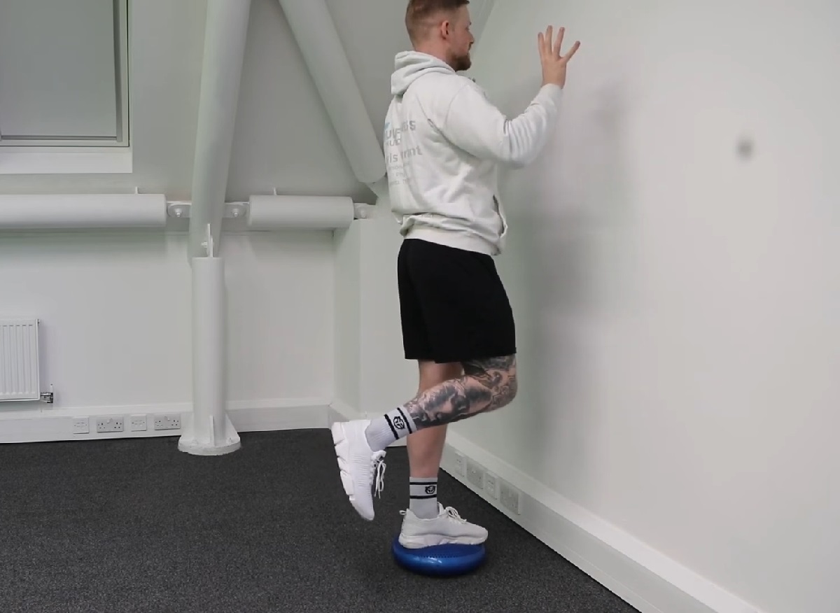 A man in a white hoodie and black shorts is balancing on one foot atop a blue stability cushion against a white wall, performing proprioceptive training for stability and injury prevention