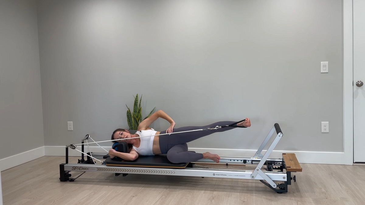 A woman performing a side-lying leg exercise on a Pilates reformer machine in a minimalist home gym