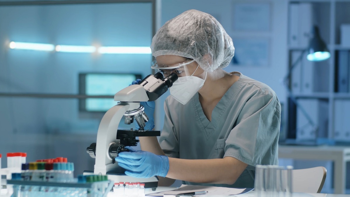 A scientist wearing protective gear examines a sample under a microscope in a laboratory