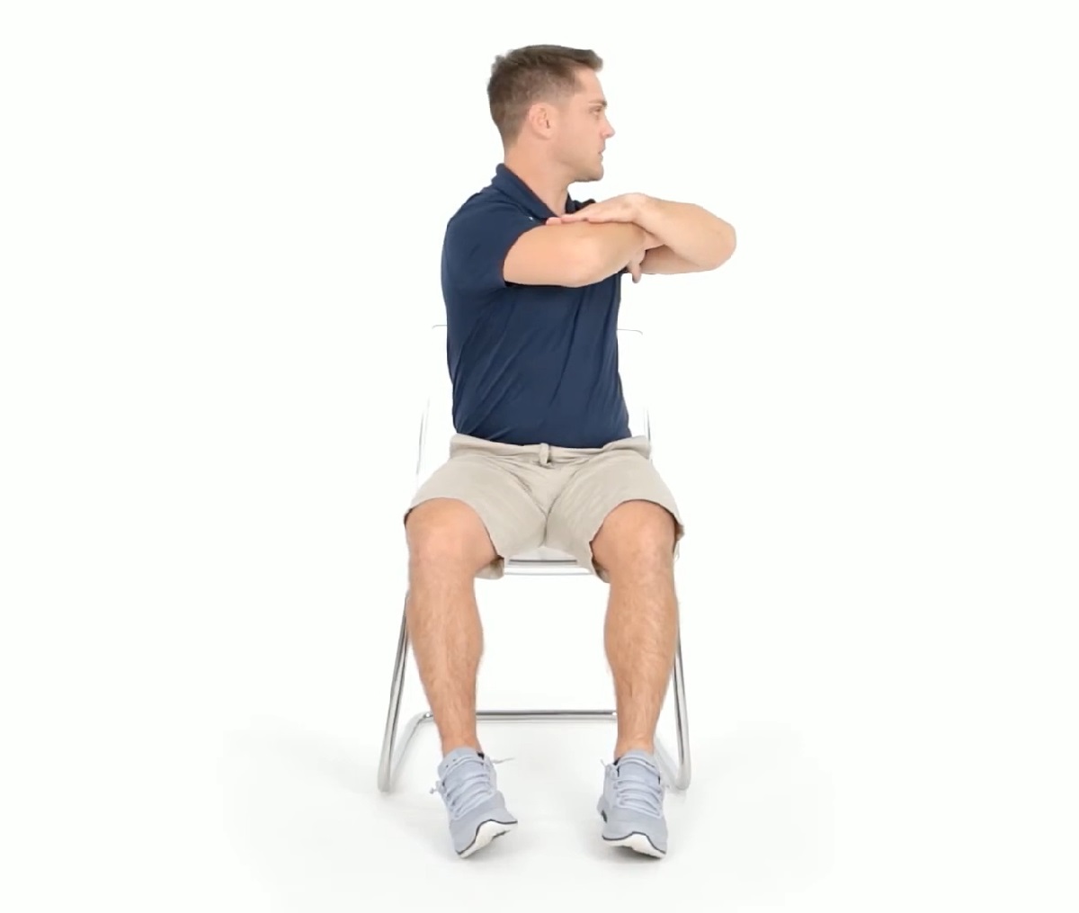 A man in a navy blue polo shirt and beige shorts sits on a white chair while twisting his torso to the side with arms crossed over his chest