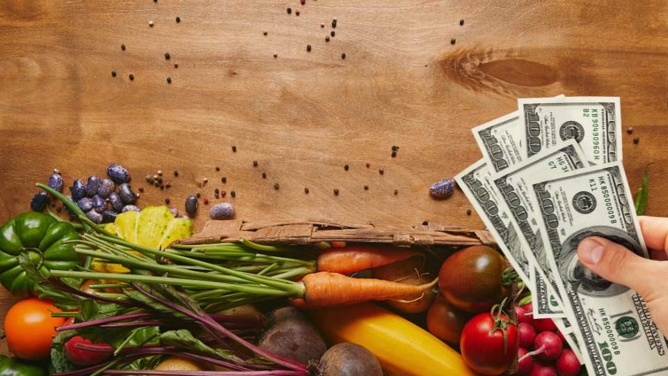 A hand holding dollar bills near a basket of colorful, fresh vegetables on a wooden surface