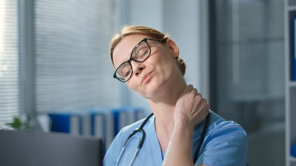 A nurse in blue scrubs stretching her neck at her workstation, promoting self-care