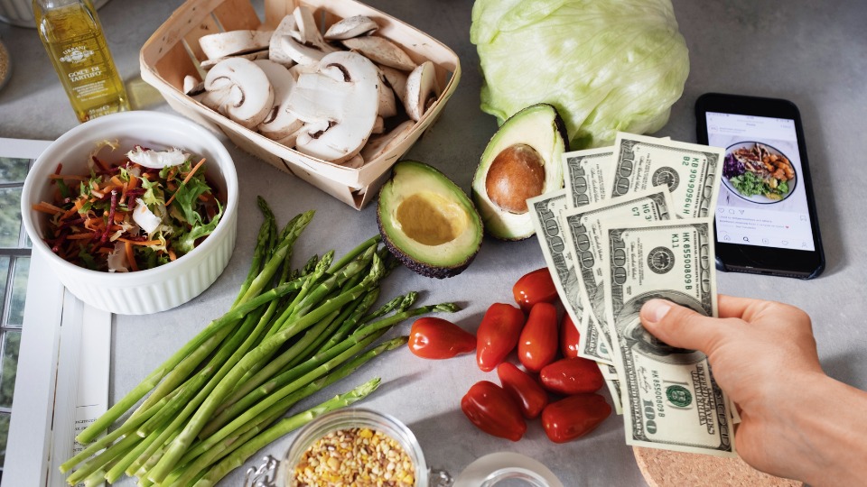 A hand holding cash near fresh vegetables, salad ingredients, and a smartphone displaying a food image