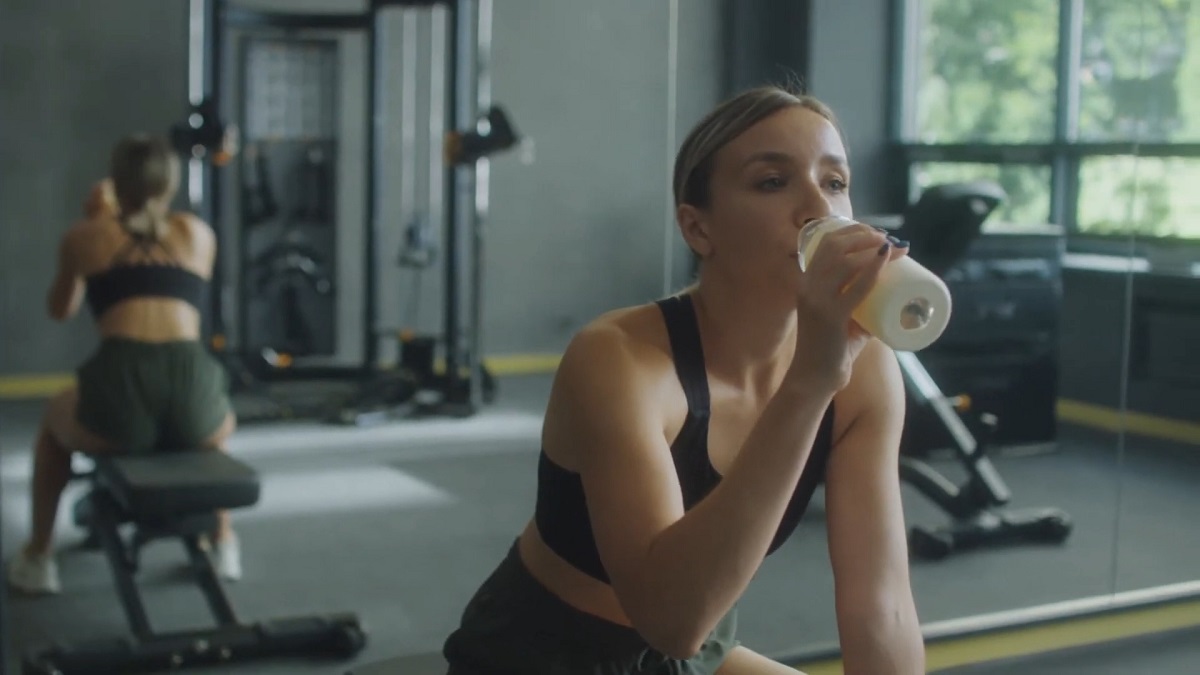 A woman in a gym drinking from a bottle post-workout while another person exercises in the background