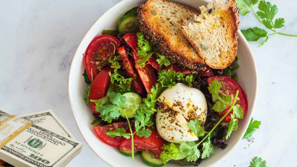 Bowl of fresh salad with tomatoes, cucumber, leafy greens, toast, and poached egg beside a stack of dollar bills
