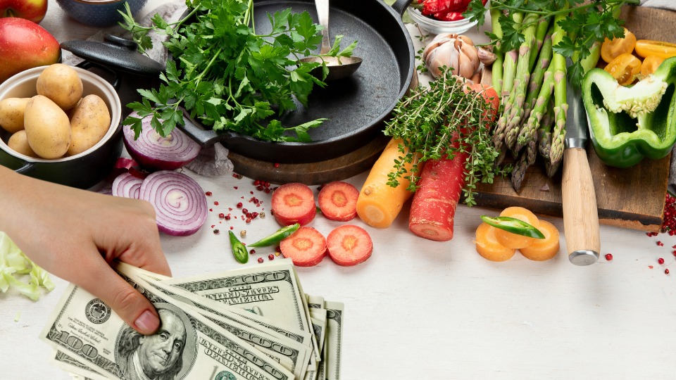 A hand holding money in front of a variety of fresh vegetables and cooking tools on a kitchen counter