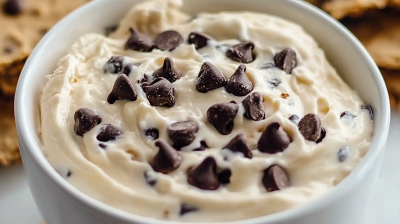 A creamy dessert dip served in a white bowl, topped with chocolate chips, resembling the flavors of a classic chocolate chip cookie