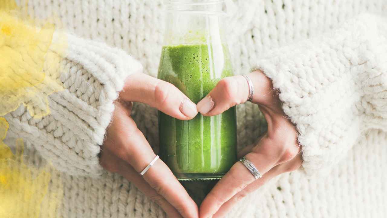 Hands forming a heart around a bottle of green juice, wearing a cozy white sweater