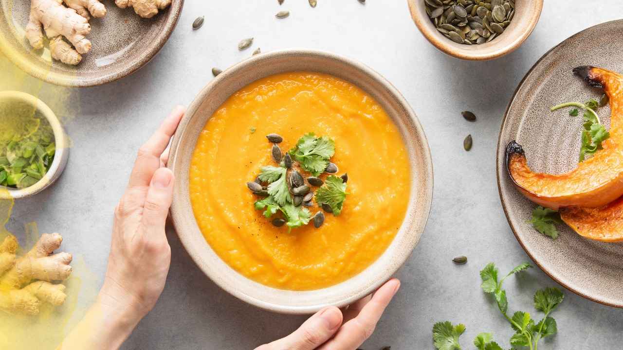 Hands holding a bowl of orange vegetable soup topped with cilantro and seeds, surrounded by fresh ingredients