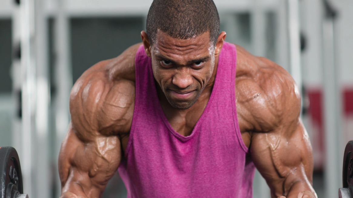 A muscular man in a pink tank top lifting weights intensely at the gym