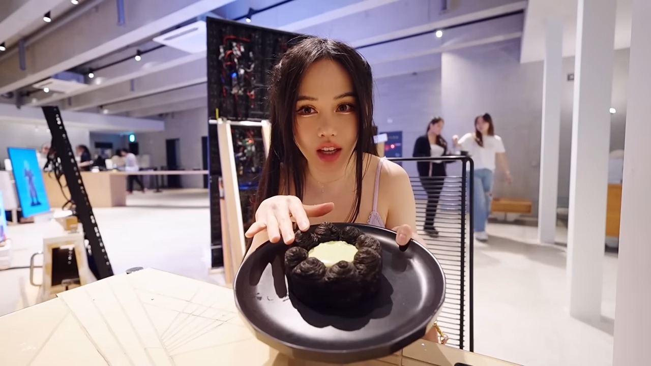 Chloe Ting, a popular fitness influencer, sitting indoors and holding a black plate with a unique dessert, smiling toward the camera.