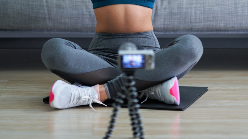 A woman in workout clothes sits cross-legged on a mat while recording fitness content with a small camera on a tripod