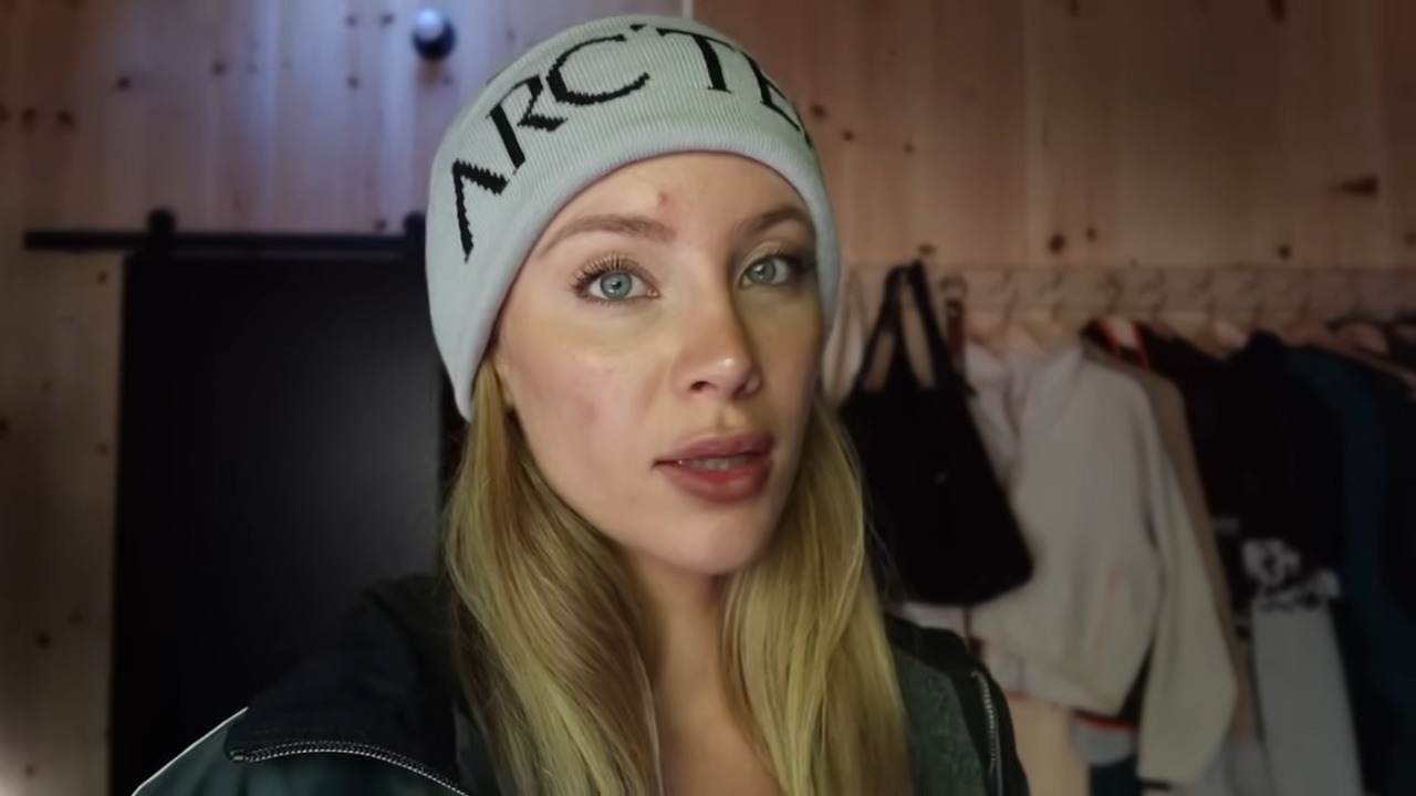 Daisy Keech, fitness model and influencer, wearing a beanie and casual jacket indoors, with coats and bags hanging in the background.