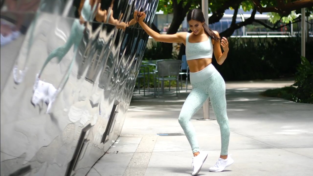 Jen Selter, fitness model and influencer, posing outdoors in a mint-green workout set with white sneakers, leaning against a reflective wall with a confident smile
