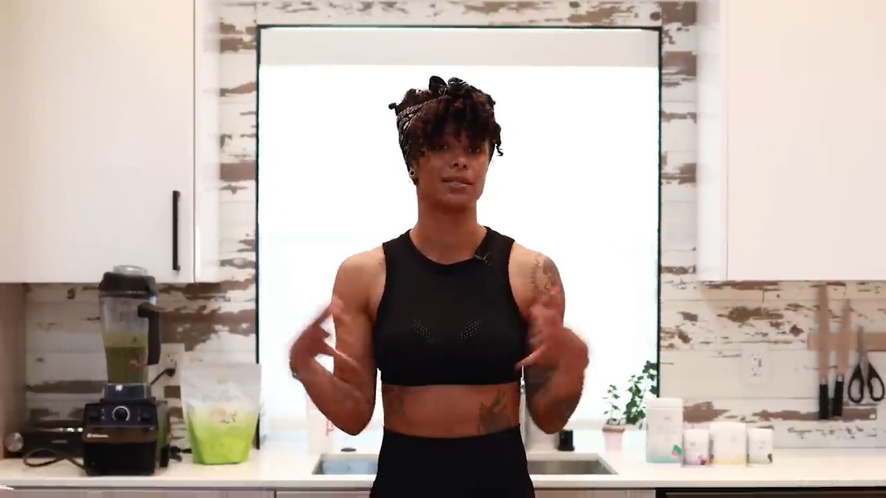 Massy Arias, fitness model and health coach, standing in a kitchen wearing a black sports bra and leggings, speaking with a blender of green smoothie behind her