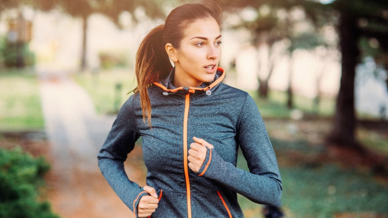 A woman jogging outdoors in a park, wearing a sporty jacket