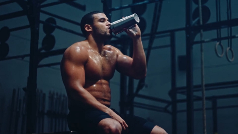 A muscular person sitting on a bench in a gym, drinking from a can