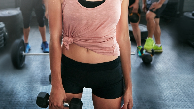 A person in a gym holds a dumbbell, with other gym-goers and weights visible in the background