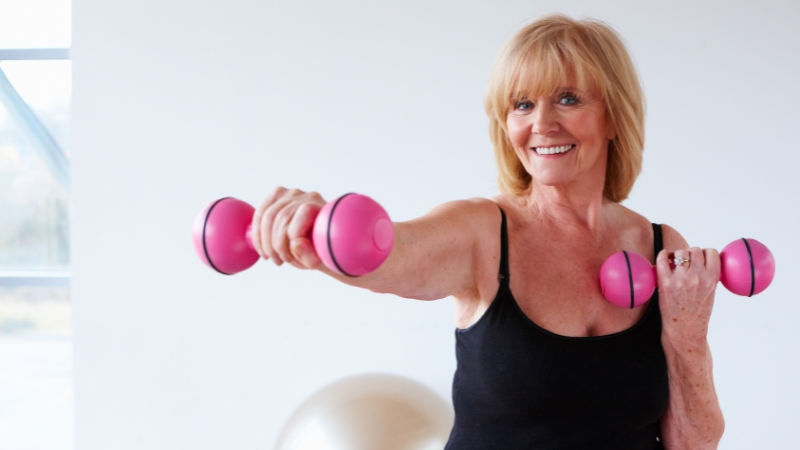 Older woman lifting weights and smiling, showing how regular exercise may help slow skin aging