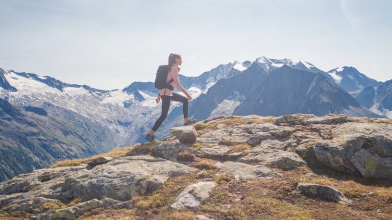 A woman hiking along a rocky ridge with snow-capped peaks in the background, prepared with gear for mountain treks