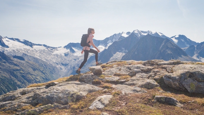 A woman hiking along a rocky ridge with snow-capped peaks in the background, prepared with gear for mountain treks
