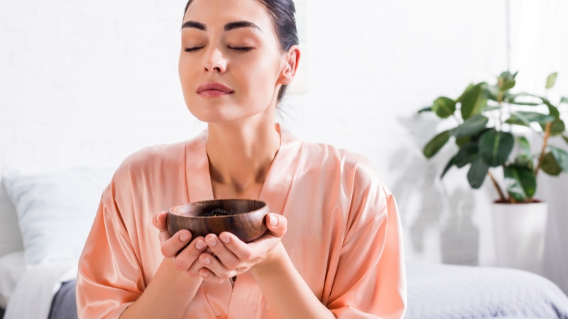 Woman in a silk robe inhaling scent from a wooden bowl during an aromatherapy session at home